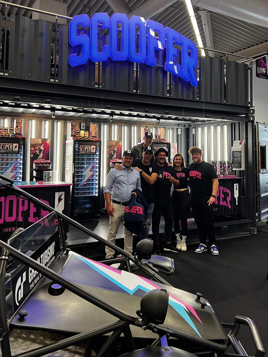 Vor dem Scooper Messestand steht ein Teil des Teams. Alle tragen Scooper Klamotten. Im Vordergrund ist der Scooper Buggy zu sehen, im Hintergrund sieht man den ausgebauten Scooper Container.