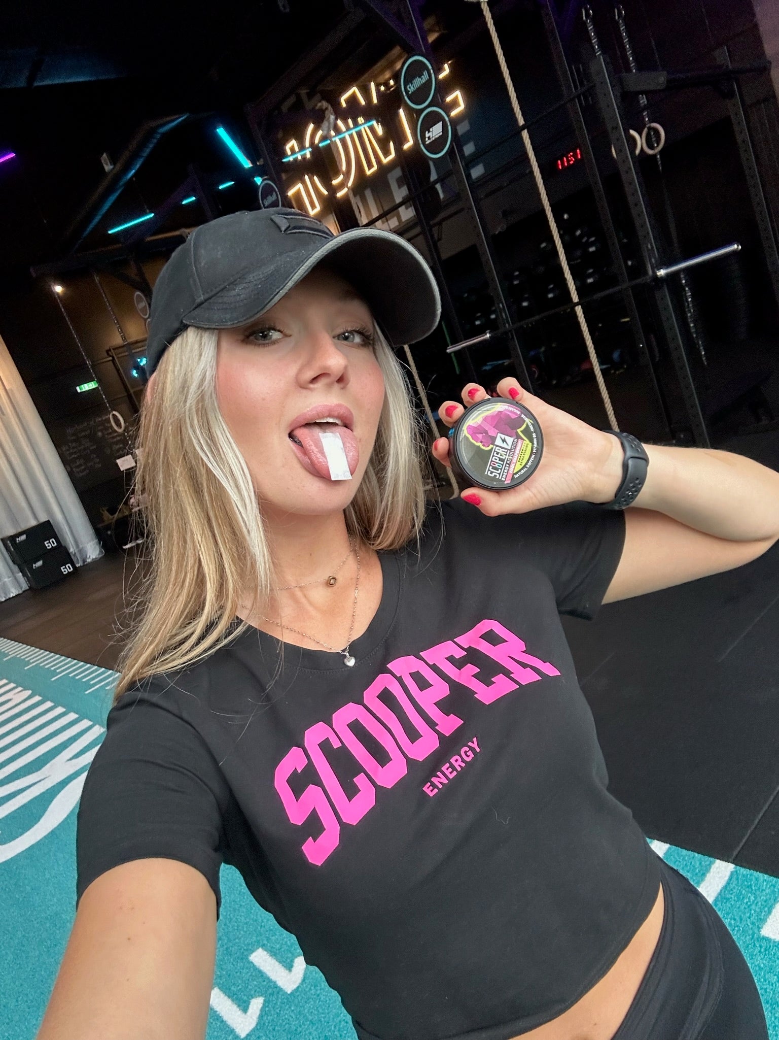 Eine Frau benutzt SCOOPER Energy im Gym. Sie steht im Vordergrund des Bildes, hält eine Dose Cool Grapefruit Extra Strong in der Hand und hat einen Pouch auf der Zunge. Die grinst mit herausgestreckter Zunge in die Camera.